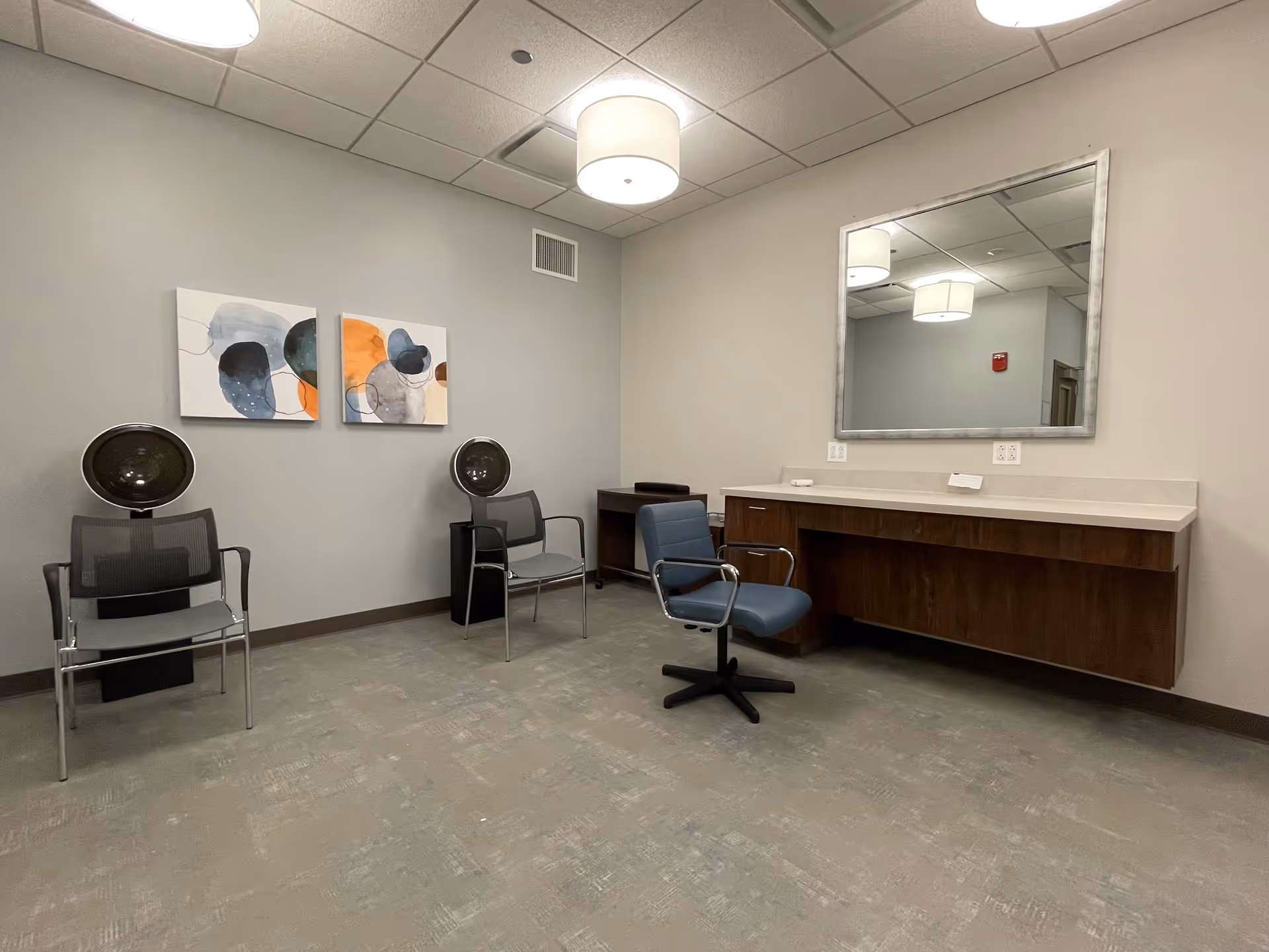 A hair salon or beauty room with two black hair dryer chairs on the left, a blue swivel chair in front of a wooden counter with a large mirror above it, and two abstract paintings on the wall. The room has gray walls, a gray carpeted floor, and ceiling lights.