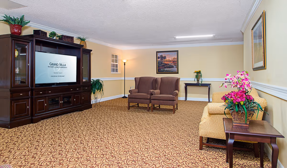 A senior living community lounge area with patterned carpet, two brown upholstered armchairs, a yellow upholstered sofa, a wooden entertainment center with a TV displaying 'Grand Villa Senior Living Community Delray East', a floor lamp, framed paintings on the walls, and decorative plants and flowers on tables and shelves.