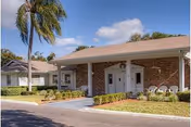 Single-story brick building entrance with a covered portico, white columns, outdoor chairs, and a palm tree nearby.