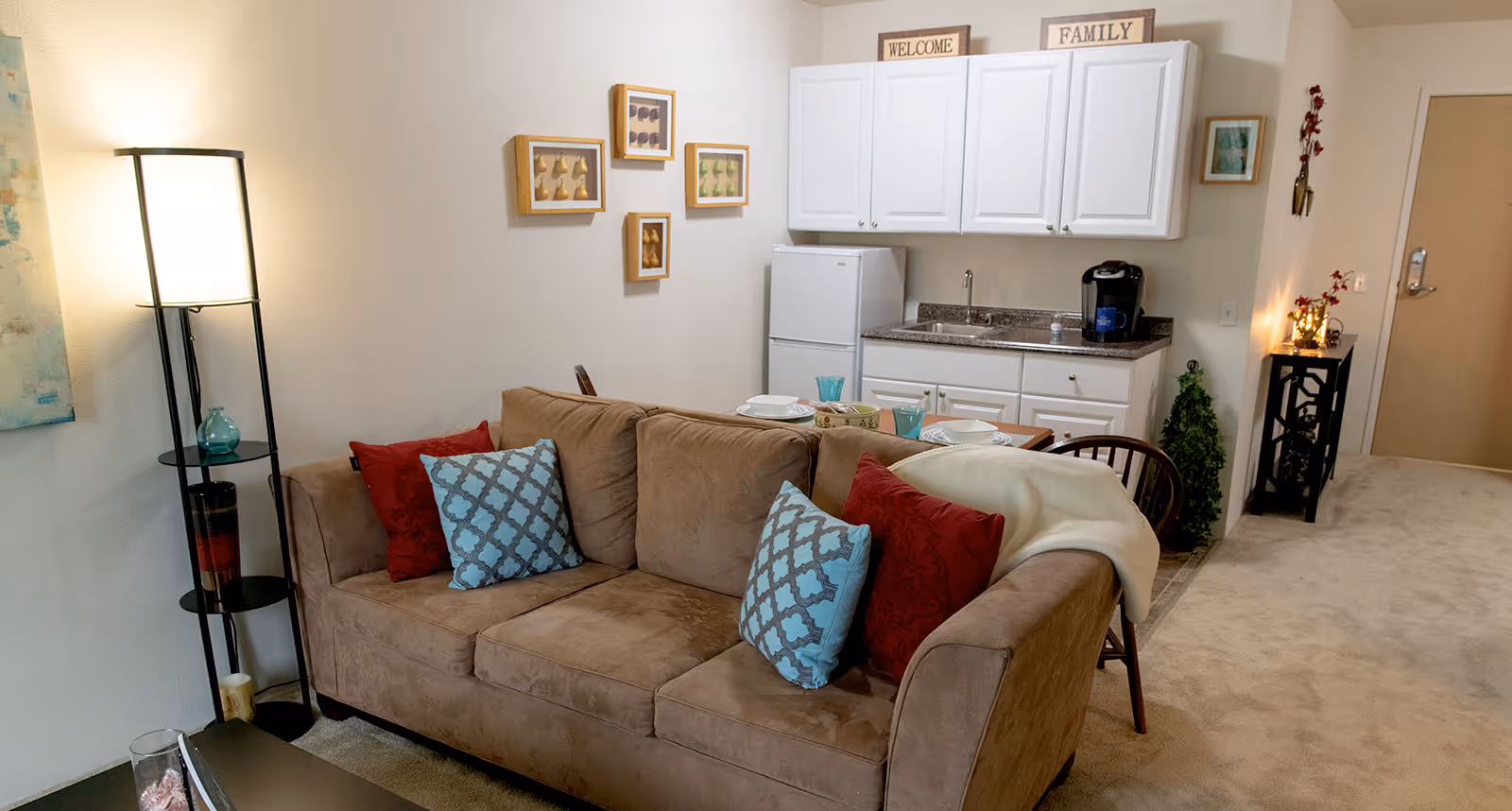 Cozy living area with a brown sofa adorned with red and blue patterned pillows, a standing lamp with shelves holding decorative items, and a small kitchen area in the background featuring white cabinets, a refrigerator, a sink, and a coffee maker. The room is decorated with framed artwork and small plants, with a carpeted floor and a door visible in the background.