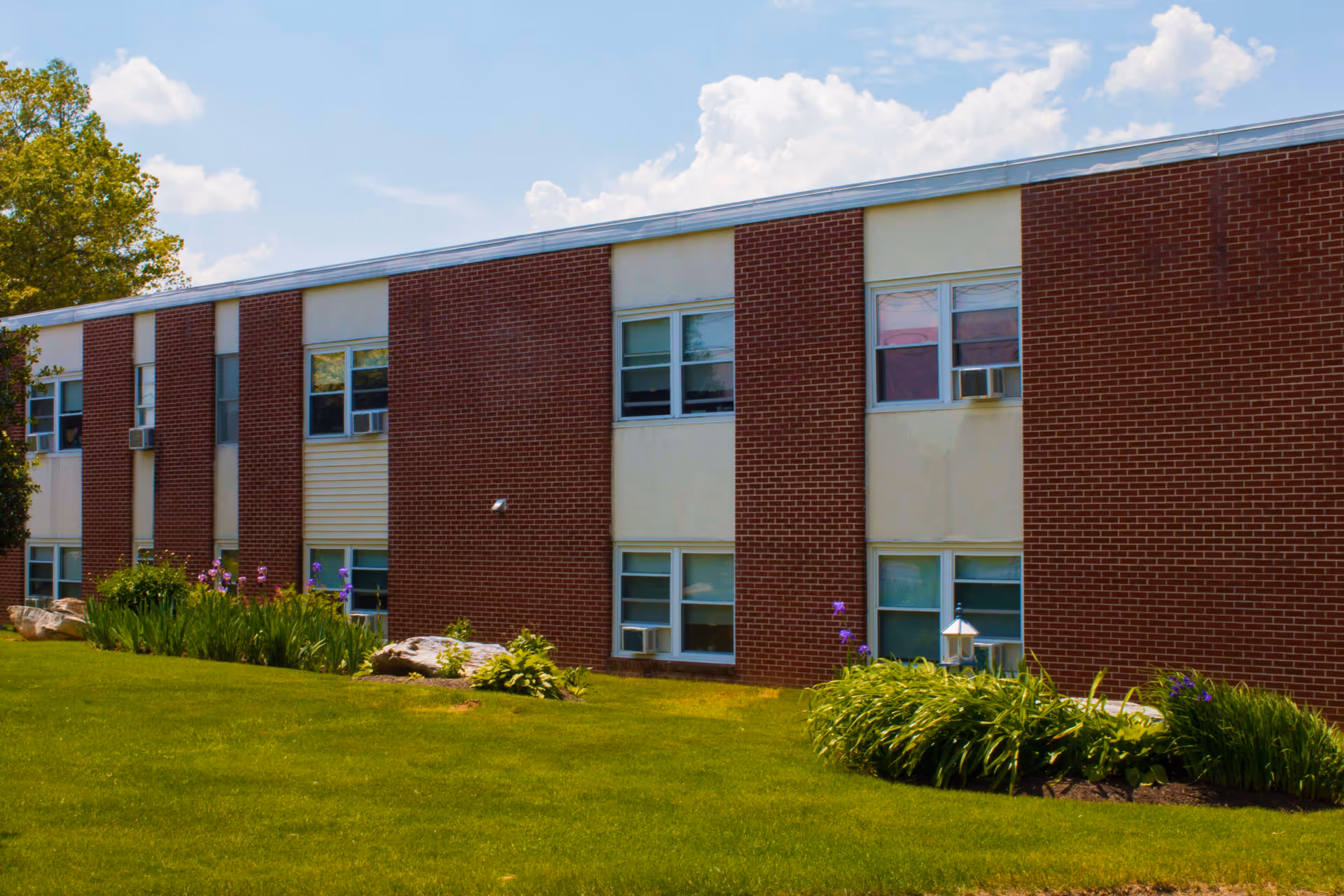 Exterior view of a two-story brick building with several windows, air conditioning units, and a well-maintained lawn with green grass and flower beds under a partly cloudy sky.