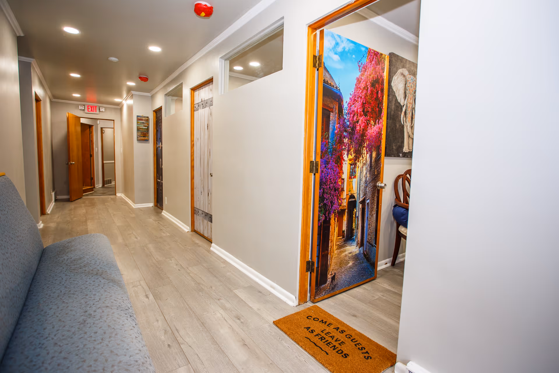 A clean, well-lit hallway in an assisted living facility with light wood flooring and beige walls. There is a blue cushioned bench along the left wall and several wooden doors on the right side. One door is open, revealing a colorful door mural of a vibrant street scene with pink flowers. A doormat in front of this door reads 'COME AS GUESTS LEAVE AS FRIENDS'. The hallway extends toward an exit door at the far end.