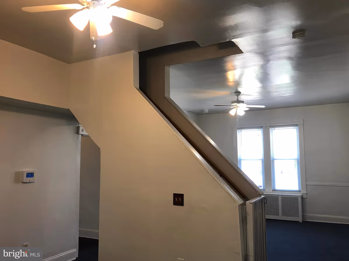 Interior view of a room with a staircase in the center, white walls, ceiling fans with lights, two windows with blinds, and dark carpeted floor.