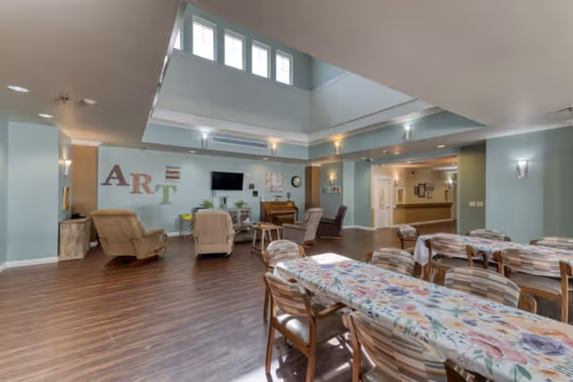 A spacious common area in a senior living facility with light blue walls and wood flooring. The room features several tables covered with floral tablecloths and surrounded by wooden chairs. There are comfortable armchairs arranged near a wall with the word 'ART' displayed, a piano, and a wall-mounted television. Large windows near the ceiling allow natural light to brighten the space.