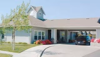 Exterior view of a single-story residential building with a covered driveway and garage. The building has light-colored siding, a small cupola on the roof, and a well-maintained lawn with a tree and flower beds.