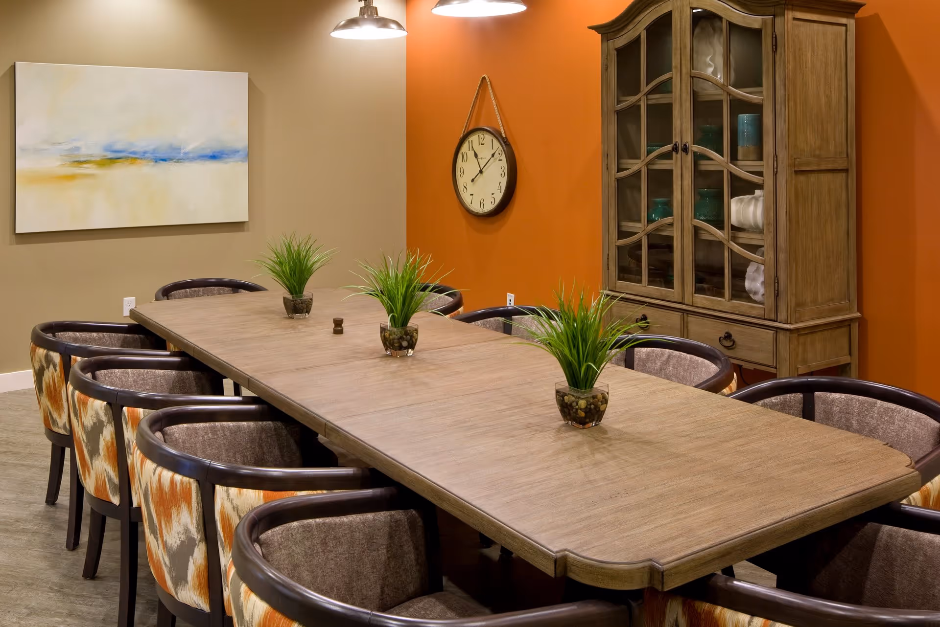 Long communal dining table with potted plants and upholstered chairs in a warmly decorated room with a wall clock and glass-front cabinet.