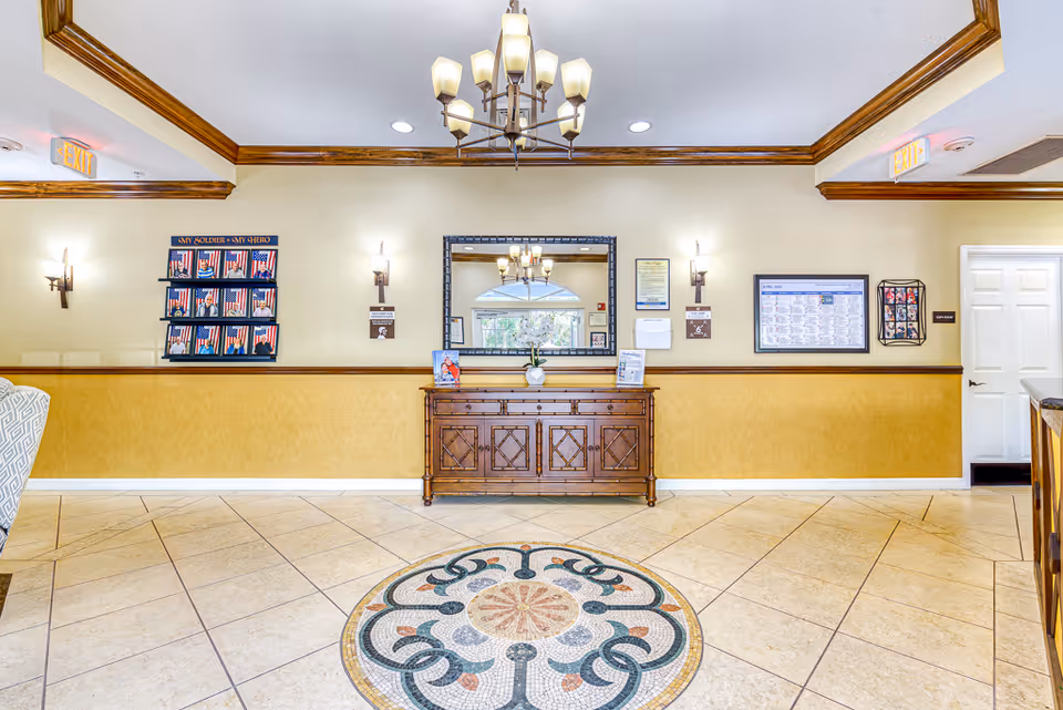 Bright senior living lobby with a decorative tiled floor medallion, wooden console beneath a mirror, chandelier, wall sconces and informational displays on the walls.