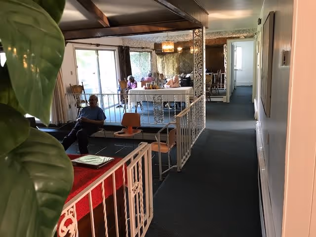 Interior view of a senior living facility showing a hallway leading to a room with a table where three elderly people are seated. The foreground includes a railing, a few chairs, and a large green plant on the left side. The room is well-lit with natural light coming through large windows.