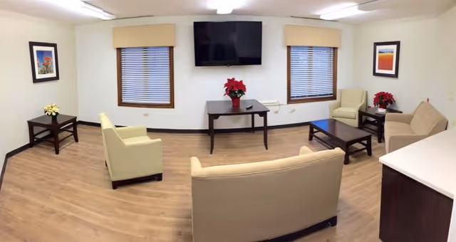 A bright communal living room with sofas, armchairs, coffee tables, a wall-mounted TV, and two windows.