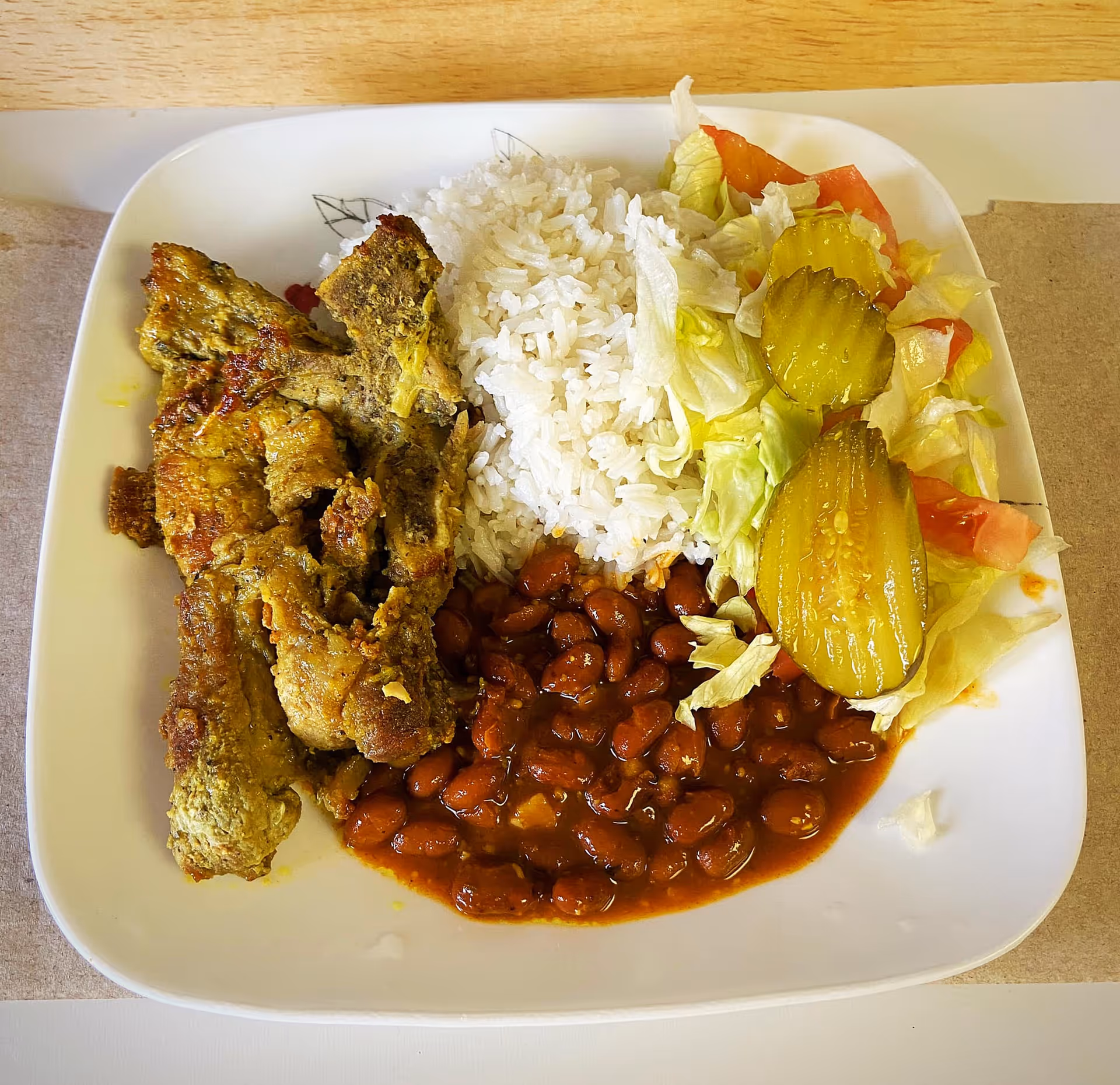 A white square plate with a serving of cooked rice, baked beans in sauce, grilled seasoned meat, and a side salad consisting of lettuce, tomato, and pickle slices on a wooden table.