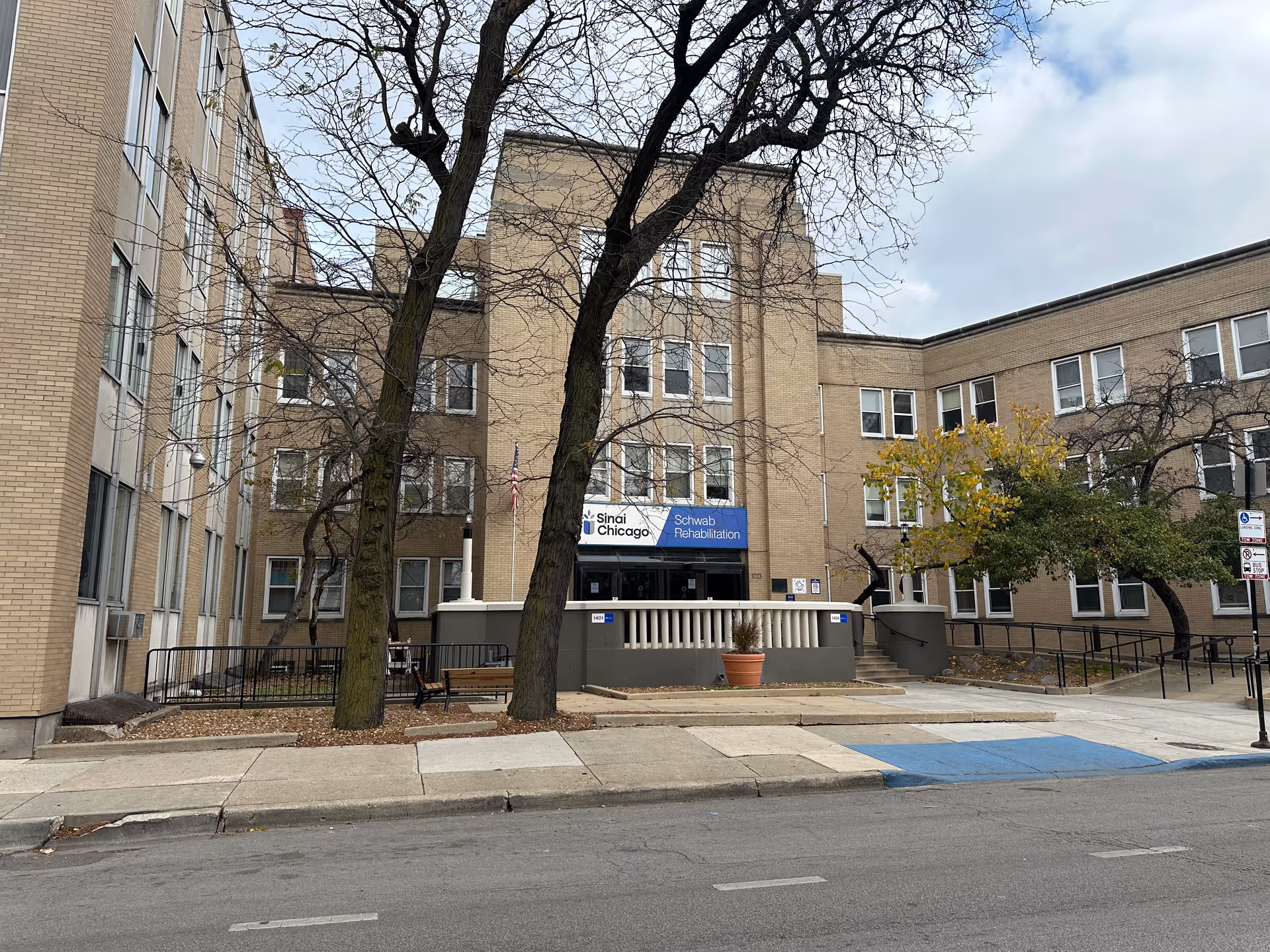 Exterior view of the Schwab Rehabilitation Center SNU building, a multi-story brick structure with several windows. There are leafless trees and some greenery in front of the building, along with a sidewalk and a street with a blue handicap parking space.