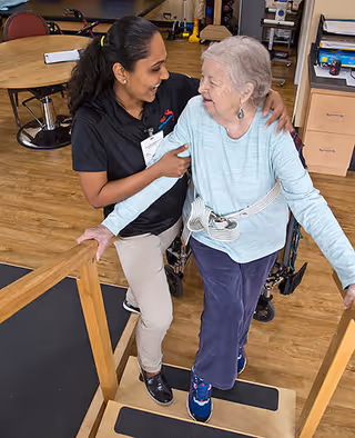 A caregiver assists an elderly woman using a walker on a wooden ramp inside a facility. The elderly woman is smiling and wearing a light blue long-sleeve shirt and dark pants, while the caregiver is smiling and supporting her with one arm around her back. The background shows tables, chairs, and office furniture on a wooden floor.