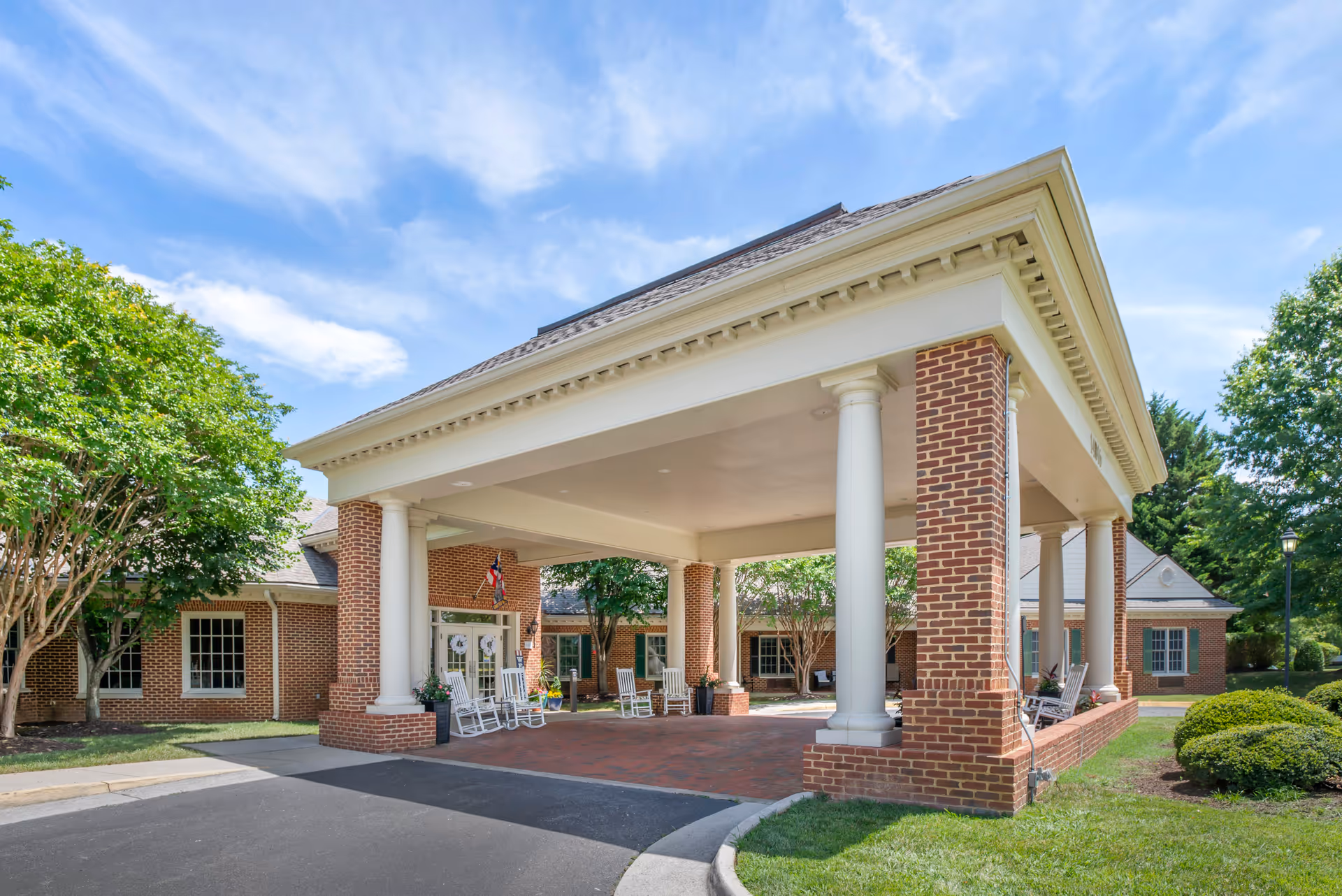 Entrance of a brick building with white columns supporting a covered driveway. Several white rocking chairs are placed under the covered area near the entrance doors. The building is surrounded by green trees and bushes under a partly cloudy blue sky.