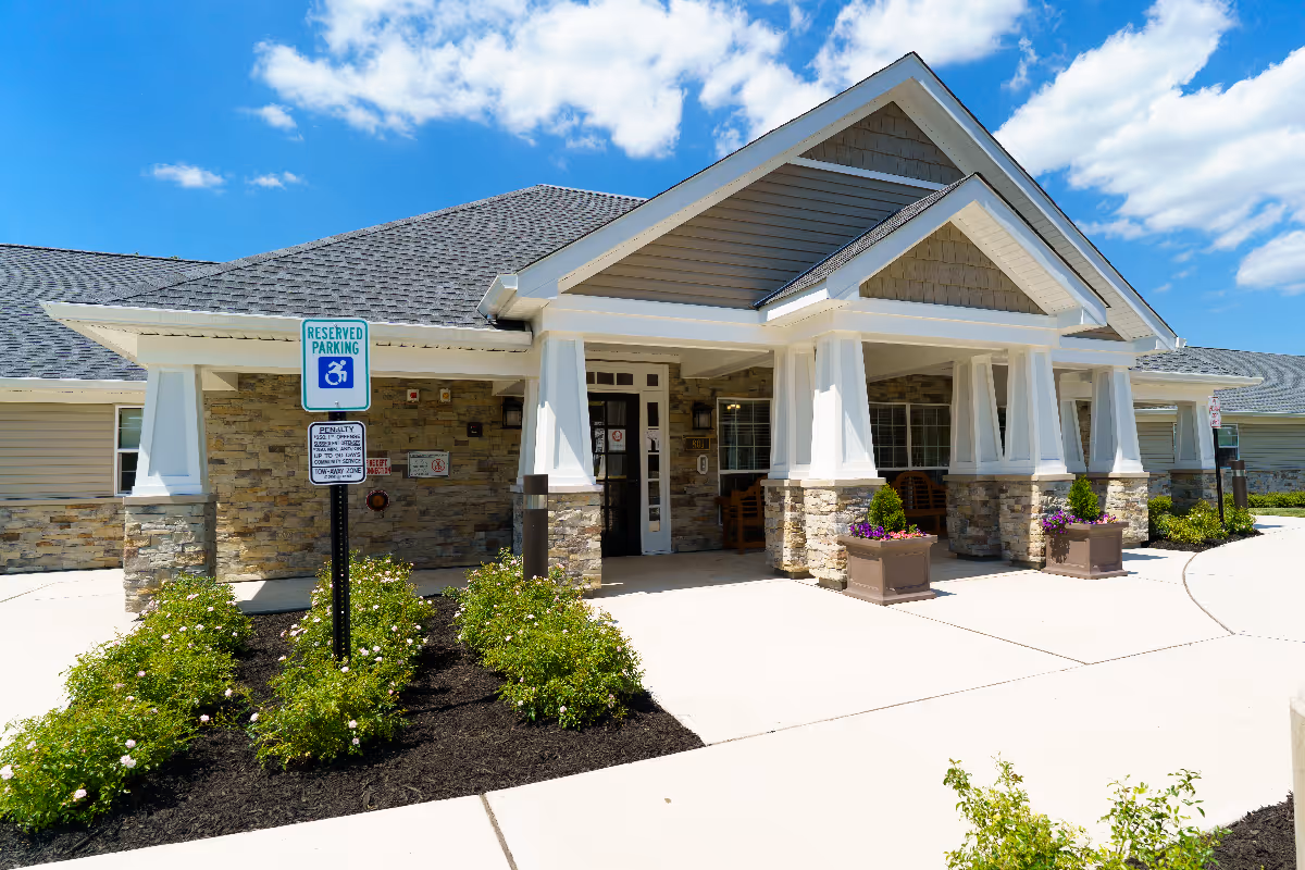 Front exterior view of a senior living facility building with a covered entrance supported by white columns and stone accents. There are flower planters near the entrance and a reserved parking sign for handicapped parking in front. The sky is blue with some clouds.