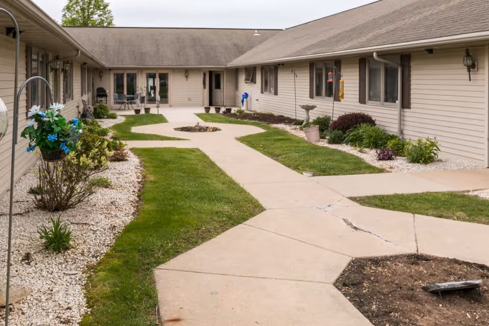 Outdoor courtyard area of Scandinavian Court Assisted Living with beige buildings surrounding a central paved walkway, green grass, flower beds with white rocks, and some outdoor seating near the building entrance.