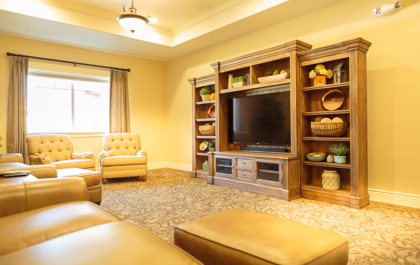 A cozy living room with beige leather sofas and two beige tufted armchairs near a window with curtains. A large wooden entertainment center with a flat-screen TV and decorative items on the shelves is against the wall. The room has warm yellow walls and patterned carpet flooring.