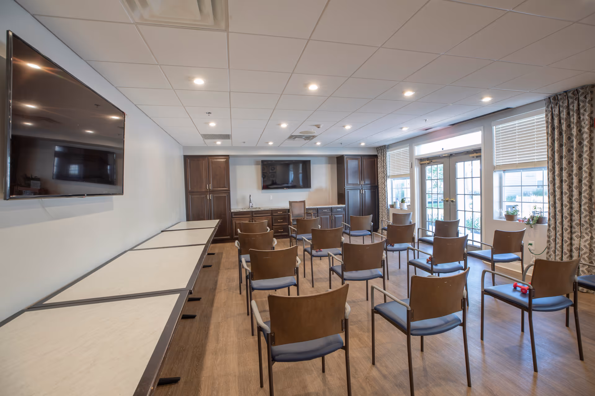 A well-lit room with multiple chairs arranged in rows facing two large wall-mounted televisions. The room has wooden flooring, white walls, and a ceiling with recessed lighting. There are large windows with patterned curtains and a door leading outside. Cabinets and a countertop are visible at the far end of the room.