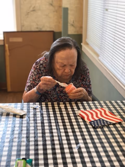 An elderly woman with long gray hair wearing a floral patterned shirt is sitting at a table with a black and white checkered tablecloth. She is eating from a small cup of pink-colored food using a spoon. On the table, there is a folded American flag napkin, a small packet, and some other items. The room has a window with blinds and light green walls.