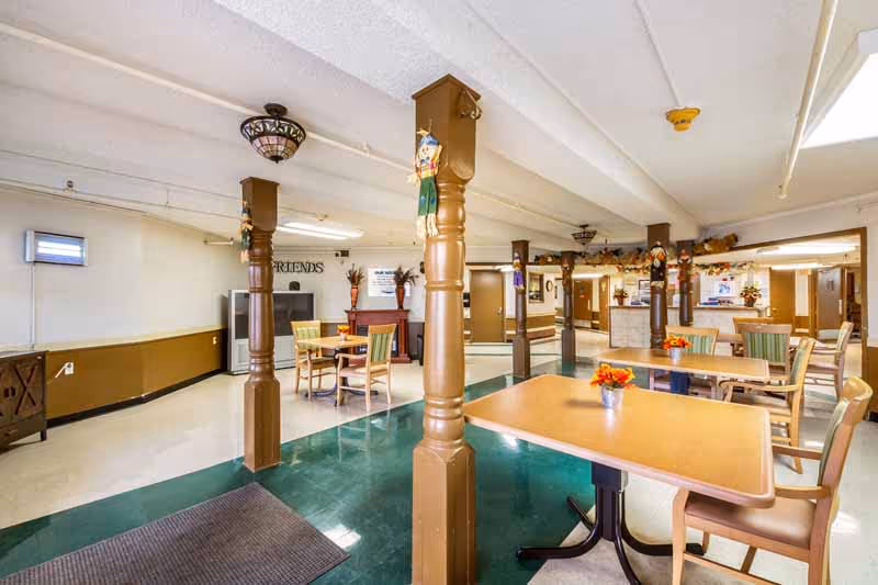 Interior view of a common area in a nursing and rehab facility featuring several tables and chairs arranged for seating. The room has brown wooden pillars decorated with small hanging ornaments, a green and white tiled floor, and a TV in the background. The walls are light-colored with brown trim, and there are ceiling lights providing illumination.