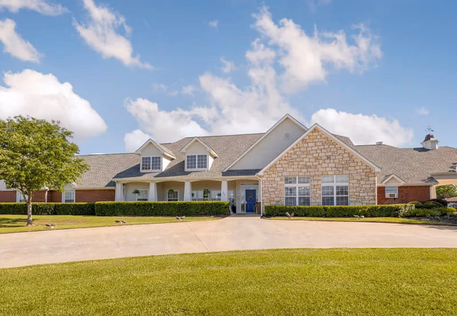 Front exterior view of a single-story senior living facility building with a combination of brick and stone facade, a blue front door, multiple windows, a well-maintained lawn, a tree on the left, and a clear blue sky with some clouds.