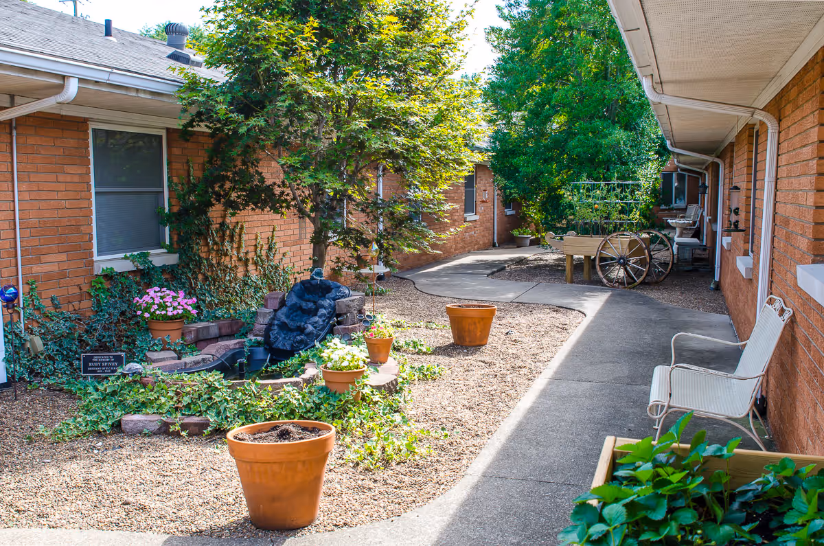 Outdoor courtyard area between two brick buildings with a paved walkway, potted plants, a small water feature, garden beds, a white metal chair, and a wooden cart with a trellis. Trees and greenery provide shade and a peaceful atmosphere.