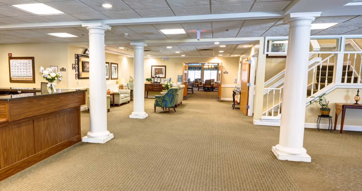 Carpeted interior lobby with white columns, a wooden reception desk to the left, seating areas and a staircase leading toward a dining area.