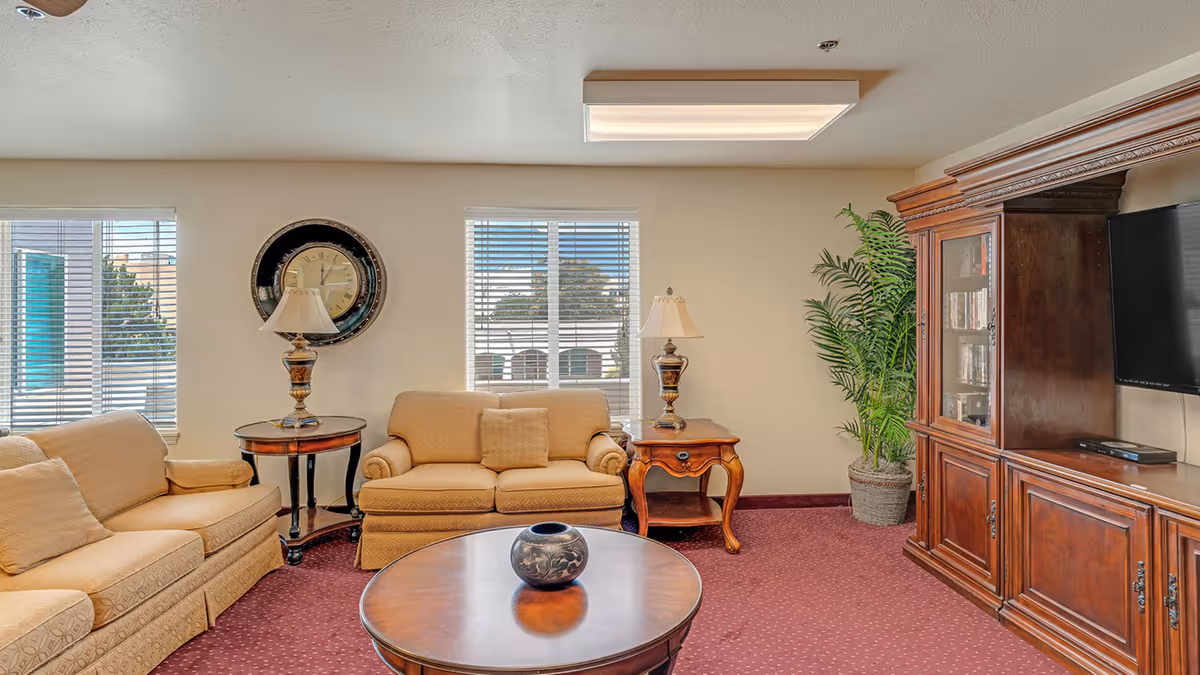 A cozy living room with two beige sofas, a round wooden coffee table with a decorative vase, two side tables with lamps, a large wooden entertainment center with a flat-screen TV, and a potted plant in the corner. The room has three windows with blinds and a maroon carpet.
