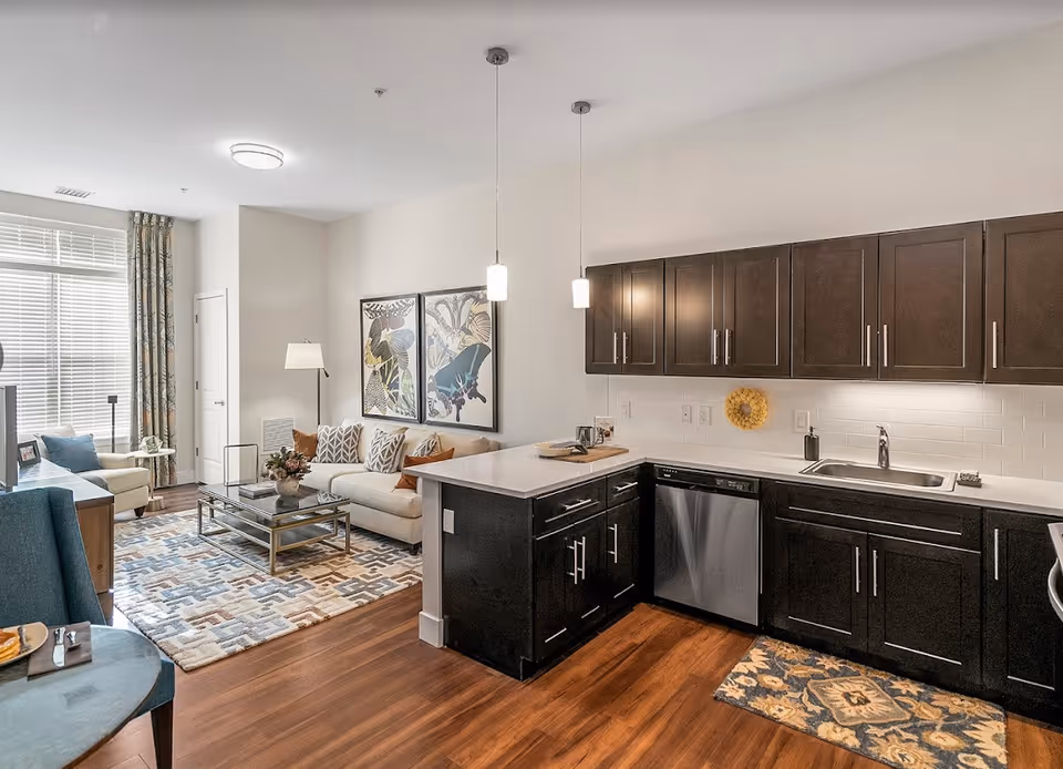 A modern open-concept living area and kitchen in a senior living facility. The living room features a beige sofa with patterned cushions, a glass coffee table on a geometric patterned rug, and a large window with curtains. The kitchen has dark wood cabinets, a white countertop, a stainless steel dishwasher, and a sink. Two pendant lights hang over the kitchen counter.
