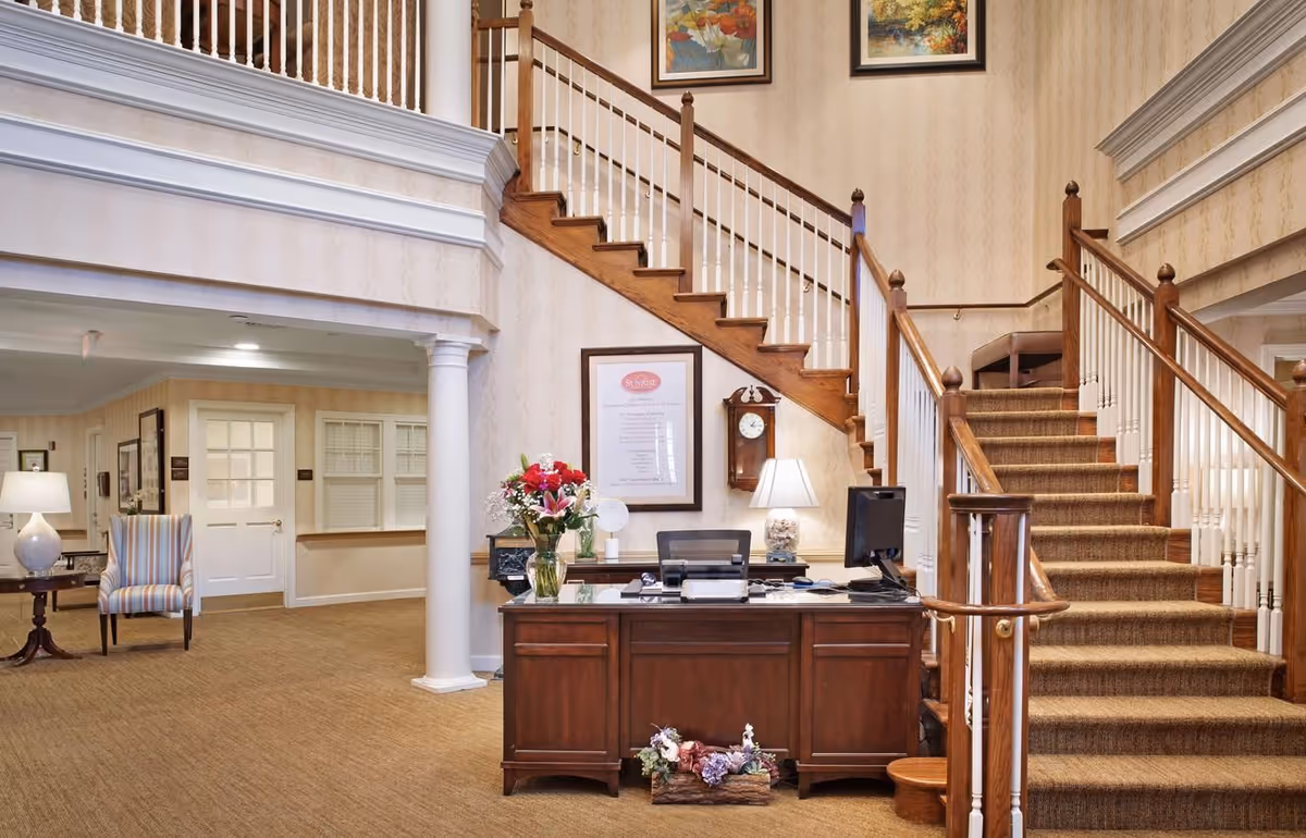 Interior view of a senior living facility reception area with a wooden desk adorned with flowers and a computer. Behind the desk is a staircase with wooden handrails and carpeted steps leading to an upper floor. The walls are decorated with framed pictures and a clock. To the left, there is a seating area with a striped armchair and a lamp on a small table.