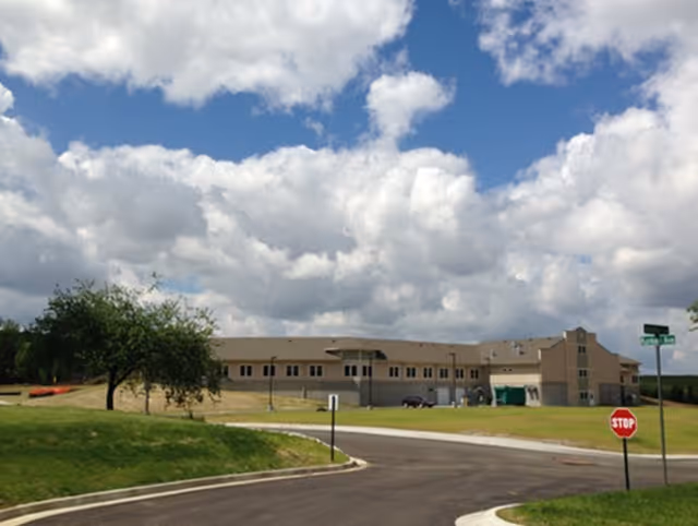 Exterior view of a single-story beige building with a pitched roof under a partly cloudy sky. The building is surrounded by grassy areas and a paved road with a stop sign and street signs visible in the foreground.