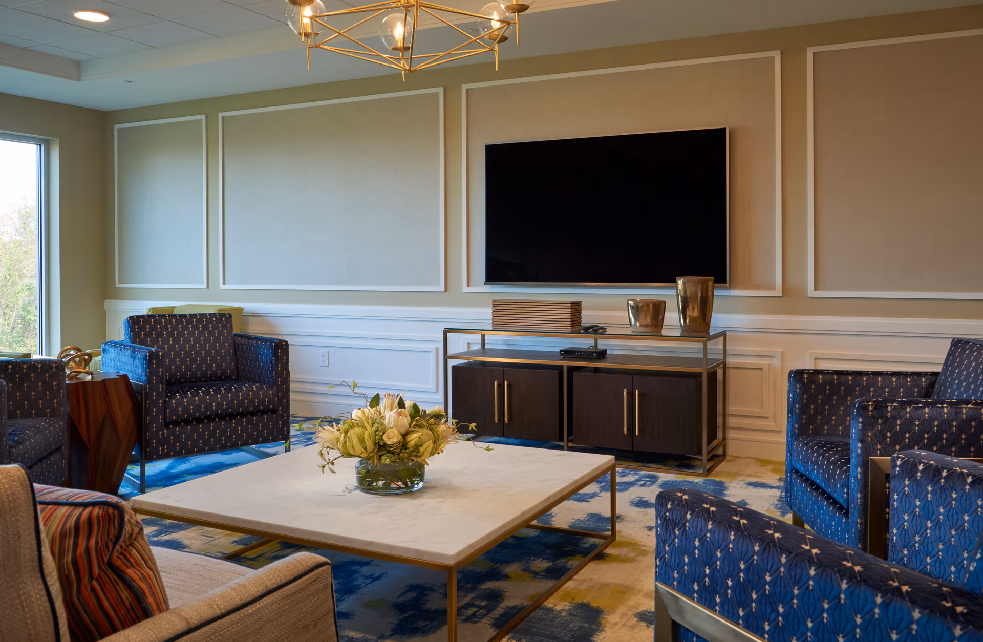 Sunlit lounge with a wall-mounted flat-screen TV, a central coffee table with a flower arrangement, and several upholstered armchairs.