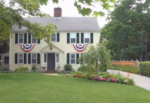 Two-story yellow house with black shutters decorated with red, white, and blue bunting on the front. The house is surrounded by a well-maintained lawn, trees, and flower beds, with a driveway on the right side.