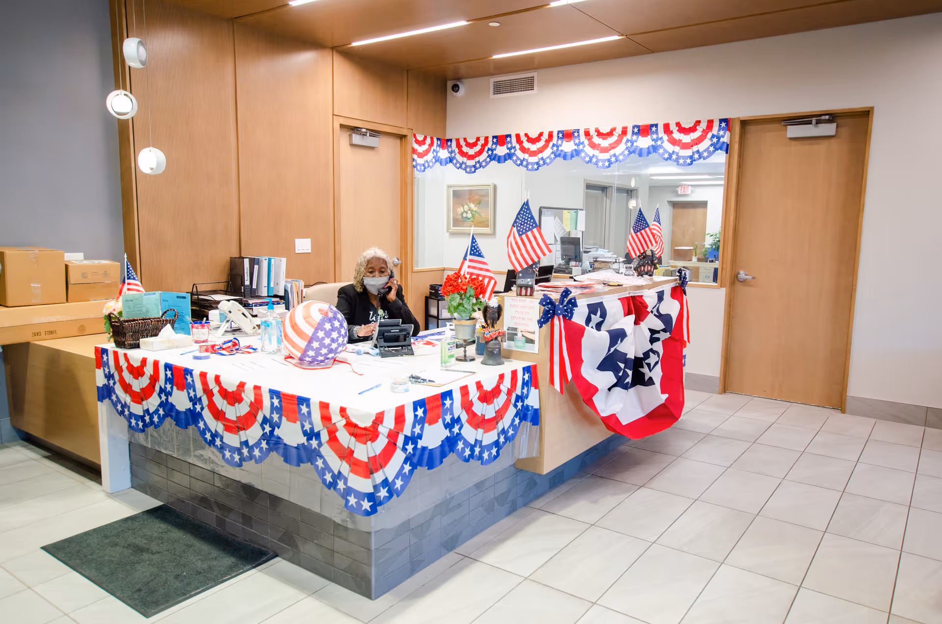 Reception desk decorated with red, white, and blue patriotic bunting and small American flags. A woman wearing a mask is sitting behind the desk, talking on the phone. The area has light-colored tiled flooring, wooden doors, and a large mirror behind the desk reflecting the room.