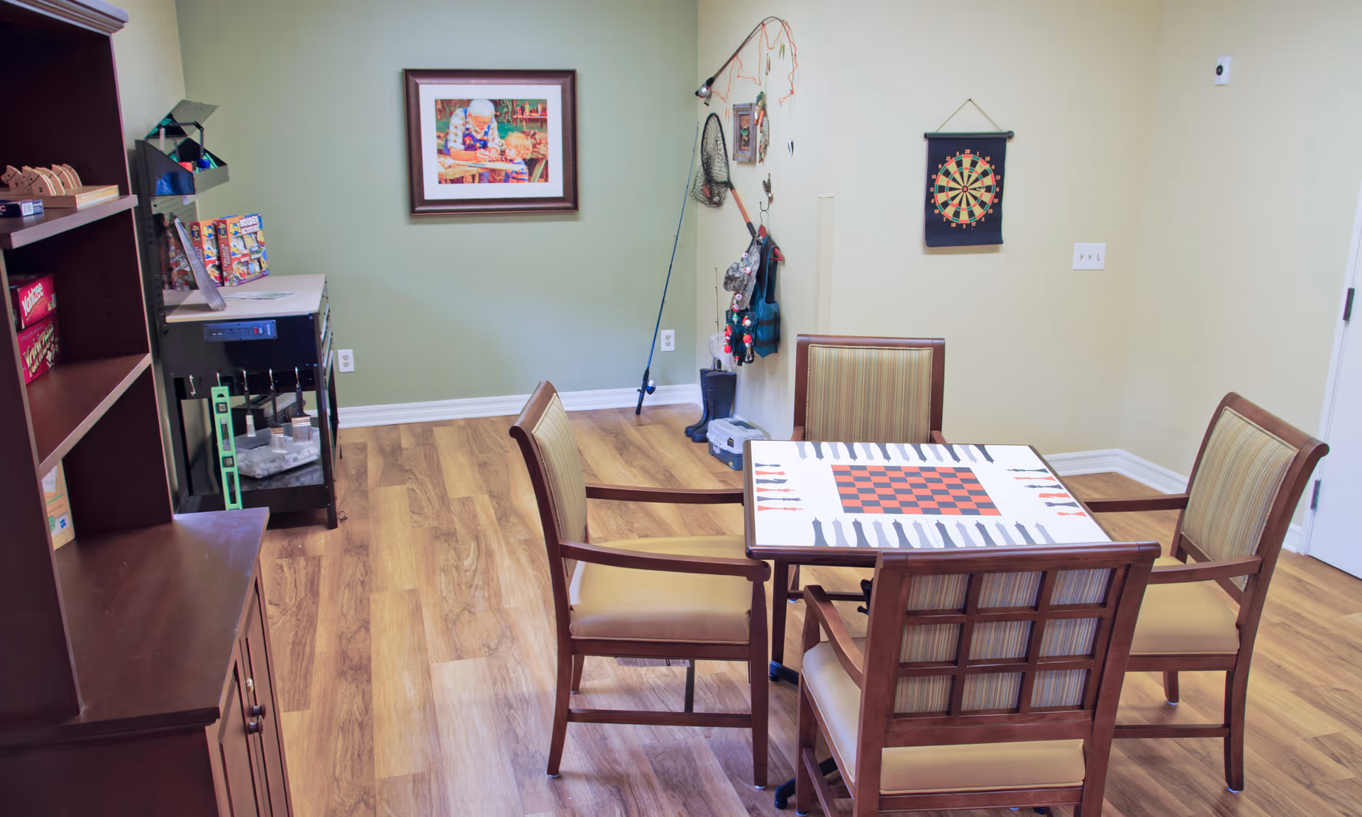 A small activity room with a square game table and four chairs, a dartboard on the wall, shelving with games, and recreational equipment against a green wall.