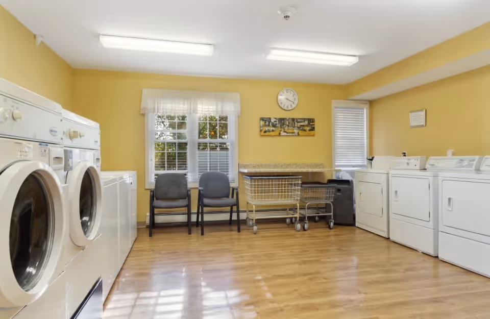 A laundry room with yellow walls and wooden flooring featuring multiple white washing machines and dryers along the left and right walls. Two gray chairs are placed in front of a window with white blinds and a sheer curtain. There are two laundry carts in the center near a countertop, and a clock and a framed picture hang on the wall above the countertop.