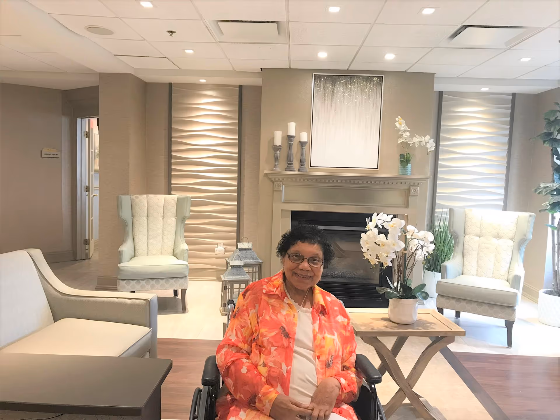 An elderly woman in a wheelchair wearing a bright orange floral shirt is smiling in a cozy living room area. The room features two light-colored armchairs, a small wooden table with a white orchid plant, a fireplace with candles and a decorative painting above it, and modern textured wall panels on either side of the fireplace.