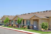Exterior view of single-story residential buildings with beige walls and brown shingle roofs, small trees and shrubs planted along the sidewalk, and a clear blue sky above.