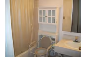 A small bathroom featuring a white sink with a mirror above it, a white cabinet with glass doors mounted on the wall, a beige shower curtain, and a beige shower chair placed in front of the bathtub.