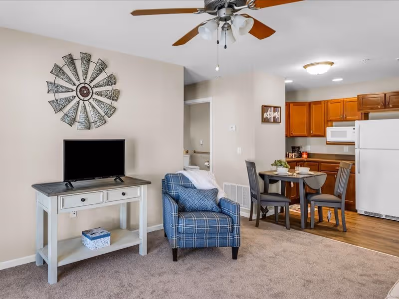 Interior view of a senior living facility unit showing a small living area with a blue plaid armchair and a TV on a white console table. The background includes a dining area with a round table and four chairs, and a kitchen with wooden cabinets, a white refrigerator, and a microwave. A decorative metal wall art piece is mounted above the TV.