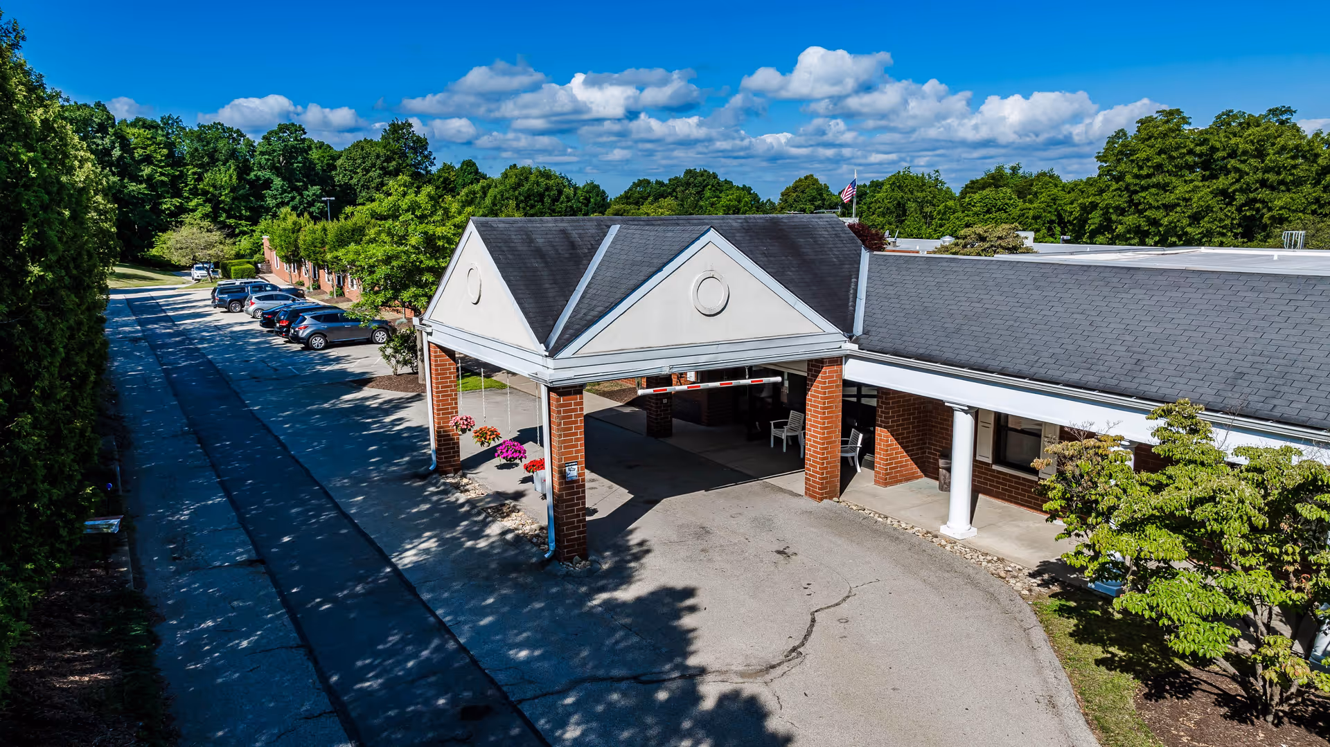 Exterior view of Kittanning Health & Rehab Center showing the main entrance with a covered drop-off area supported by brick columns and white pillars. Several cars are parked along the driveway lined with trees and greenery under a blue sky with scattered clouds.