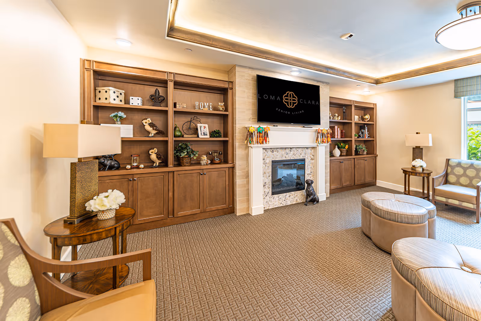 Comfortable senior living lounge with built-in wooden shelving, a fireplace and TV, and upholstered seating with ottomans.