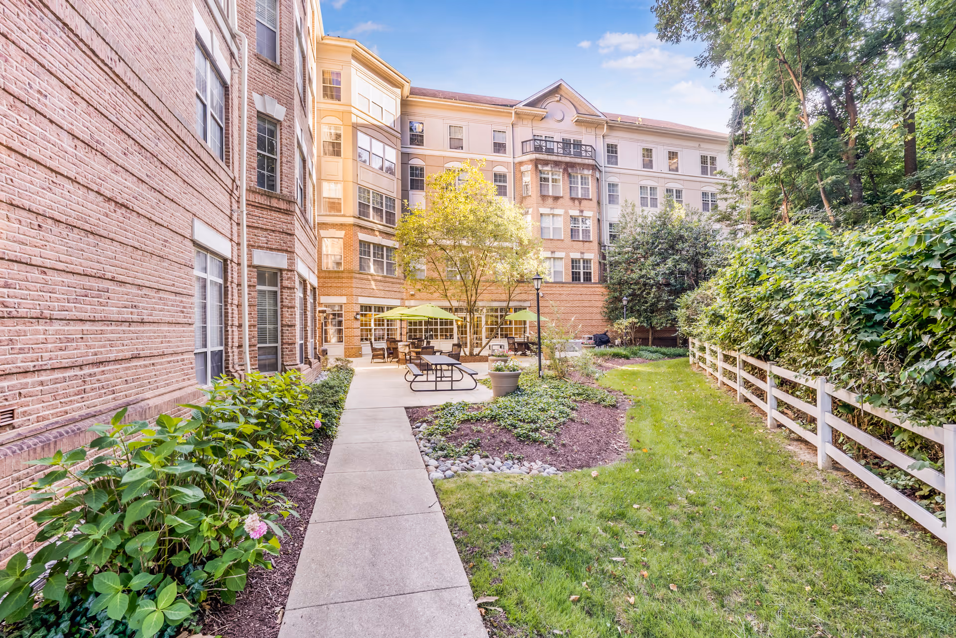 Well-maintained courtyard with a paved walkway, patio tables and umbrellas, landscaped beds and a multi-story brick building in the background.