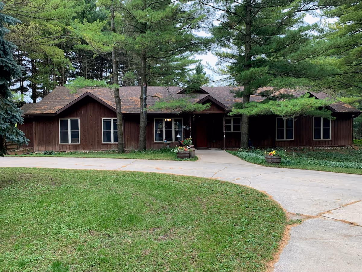 Single-story brown wooden lodge surrounded by trees with a curved driveway and flower planters at the entrance.