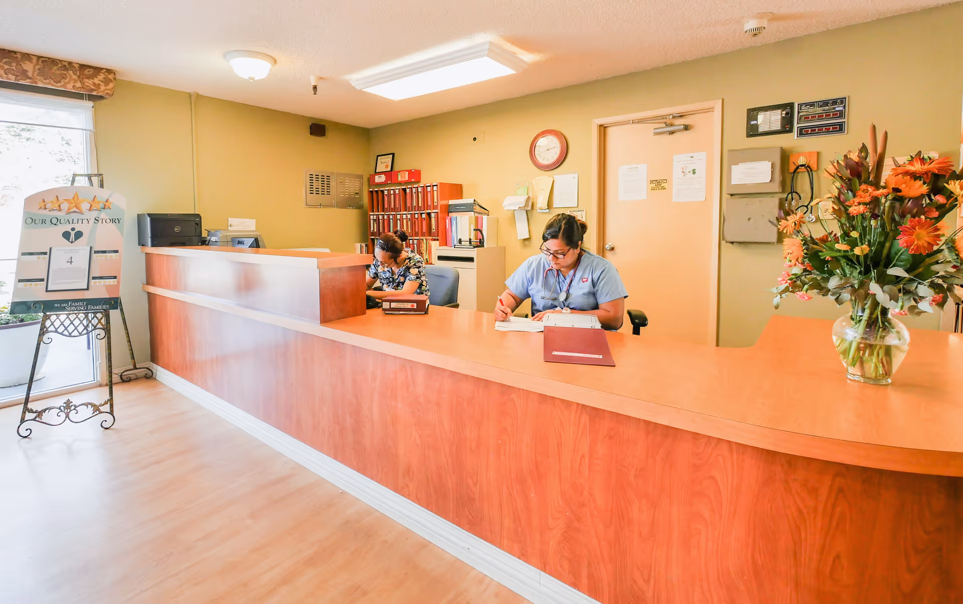 Reception area of a skilled nursing and subacute center with a long wooden front desk. Two staff members are seated behind the desk, one writing in a book and the other looking down. There is a large bouquet of orange flowers on the right side of the desk. On the left side near the entrance, there is a stand with a sign titled 'Our Quality Story'. The walls are light yellow, and there is a clock and various notices on the wall behind the desk.
