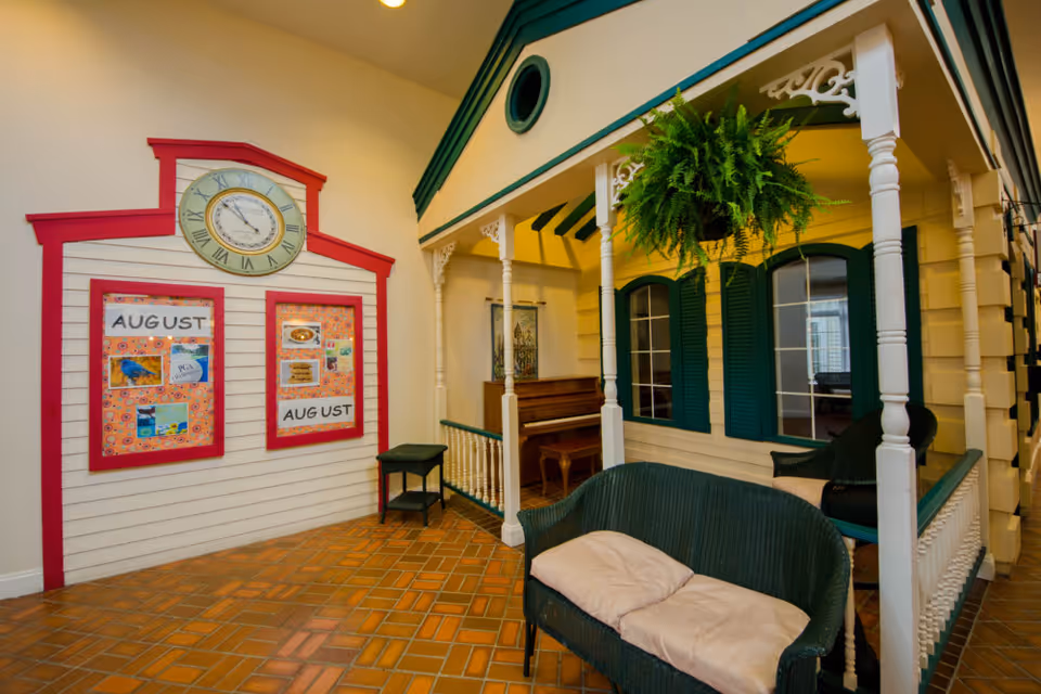 Indoor common area styled like a small house porch with wicker seating, a hanging fern, a piano and a bulletin board labeled "August".