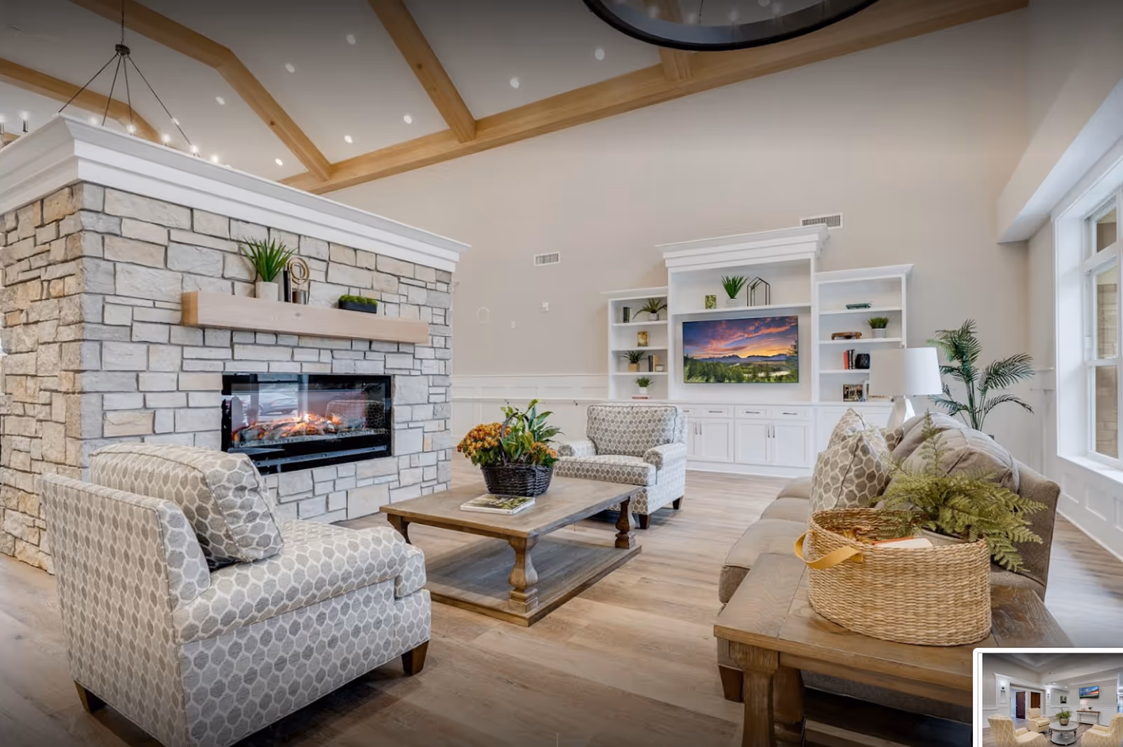 A bright and spacious living room with a stone fireplace in the center, surrounded by comfortable patterned armchairs and a sofa. A wooden coffee table with a plant centerpiece sits in the middle. The room features a high ceiling with wooden beams, large windows letting in natural light, and white built-in shelves with a TV displaying a scenic sunset.