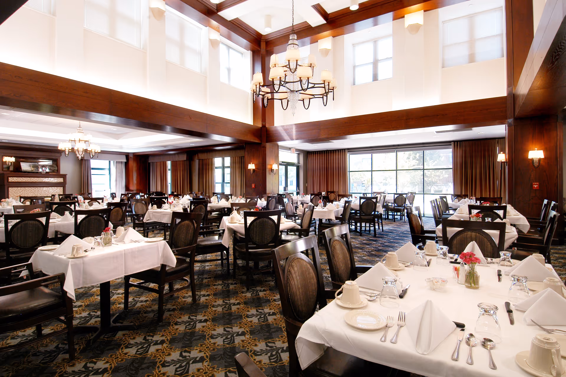A spacious, elegant dining room with multiple tables covered in white tablecloths, each set with plates, cups, silverware, and napkins. The room features large windows with brown curtains, high ceilings with wooden beams, and chandeliers providing warm lighting.