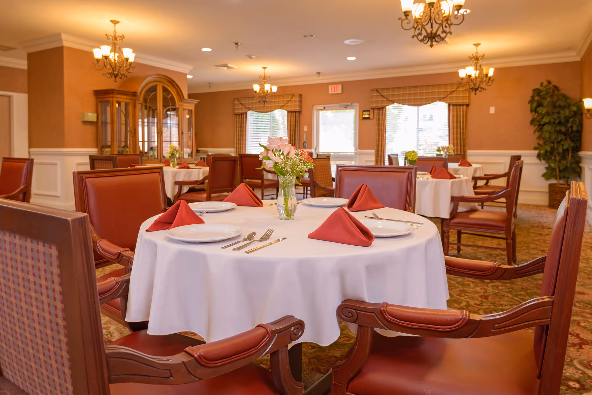 Elegant dining room with round tables set with white tablecloths, red napkins, place settings and chandeliers.