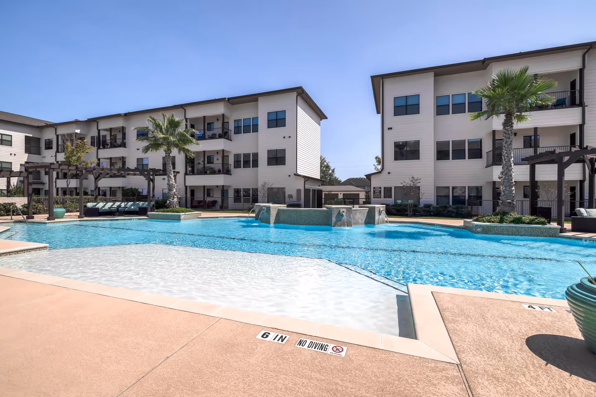 Outdoor swimming pool area with clear blue water, surrounded by a beige concrete deck. Two three-story white residential buildings with balconies and palm trees are visible in the background under a clear blue sky.