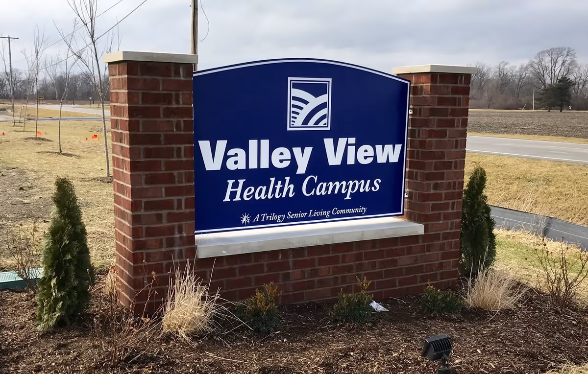 A large blue sign with white text that reads 'Valley View Health Campus, A Trilogy Senior Living Community' mounted on a brick base with two brick pillars on either side, situated outdoors near a road with some small trees and grass around it under a cloudy sky.